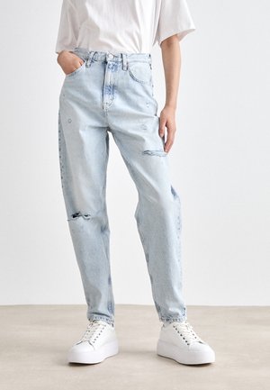 Persoon met lichtblauwe gescheurde spijkerbroek en witte sneakers, staand met één hand in de zak tegen een effen achtergrond.