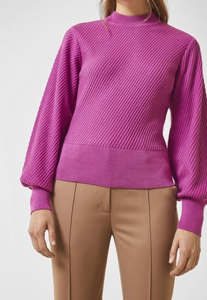 Femme portant un pull magenta côtelé à manches longues avec des manches bouffantes et un pantalon beige taille haute sur un fond uni.