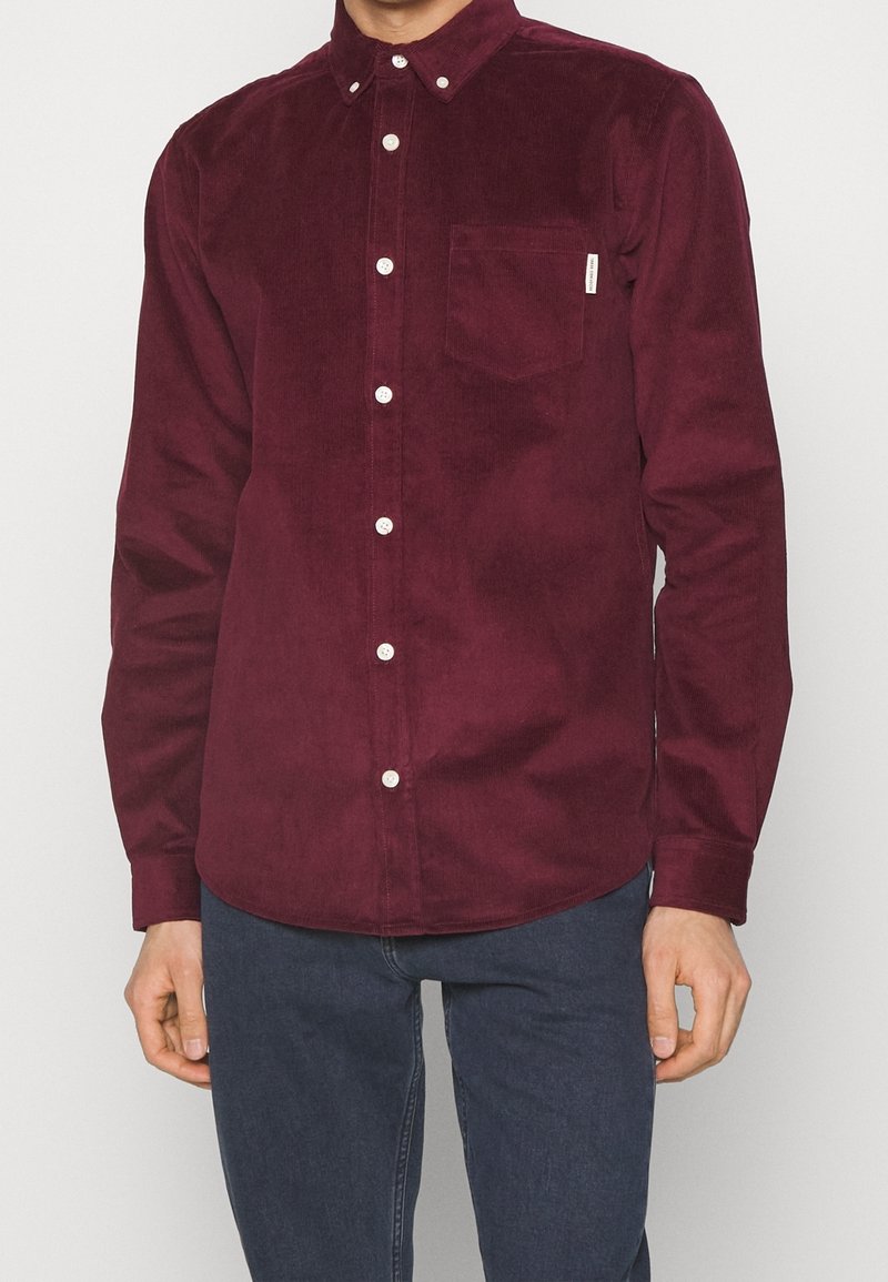 Homme portant une chemise en velours côtelé rouge foncé à manches longues boutonnée avec une poche poitrine et un pantalon bleu foncé.