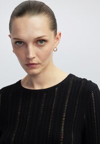 Schwarzer Strickpullover mit vertikalen Texturlinien, rundem Ausschnitt und gerippten Bündchen. Das Modell trägt goldene Creolen und hat eine gepflegte Frisur.