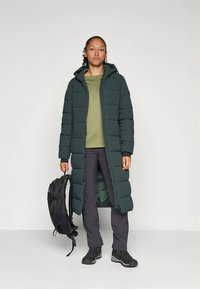 Lang, mørkegrøn pufferjakke med hætte, der har et quiltet design. Kombineret med en grøn top, mørke bukser og sorte støvler, holder en rygsæk.