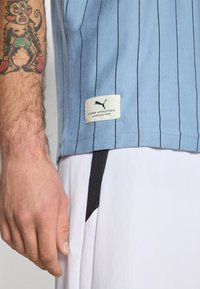 Lichtblauwe sportshirt met verticale zwarte strepen, voorzien van een wit Puma-label aan de onderkant; gedragen met witte shorts en een zwarte koord.