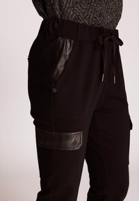 Schwarze Joggerhose mit Kordelzug am Bund, Lederakzenten an den Taschen, glatter Textur und einem Cargo-ähnlichen Patch am Oberschenkel.