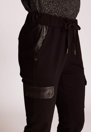Schwarze Joggerhose mit Kordelzug am Bund, Lederakzenten an den Taschen, glatter Textur und einem Cargo-ähnlichen Patch am Oberschenkel.