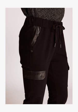 Black jogger pants featuring a drawstring waist, leather accents on pockets, smooth texture, and cargo-style patch on the thigh.