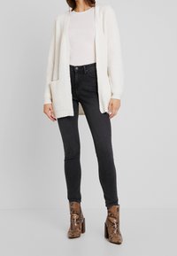 Biała dziergana kardigan z otwartym przodem i kieszeniami, zestawiony z jasną bluzką i czarnymi obcisłymi dżinsami, uzupełniony brązowymi botkami w wężowy nadruk.