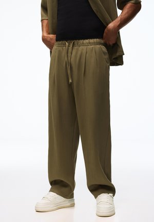 Osoba ubrana w oliwkowozielone, szerokie spodnie z sznurkiem w pasie, czarną koszulę, oliwkowozieloną otwartą koszulę i białe trampki, ręce w kieszeniach.