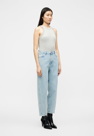 Μια γυναίκα στέκεται με το πρόσωπό της προς τα εμπρός, φορώντας ένα γκρι αμάνικο τοπ, ανοιχτόχρωμα τζιν ψηλόμεσα και μαύρες μυτερές μπότες με ψηλό τακούνι σε λευκό φόντο.