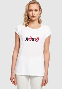 Valkoinen puuvillapaita, jossa käännetyt hihat, esittelee punasävyisen ja mustan "XOXO" -grafiikan keskellä. Paidassa on sileä rakenne ja rento muotoilu.