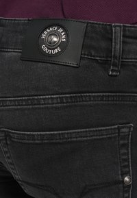 Artimas juodos Versace Jeans Couture etiketės ir nugarinės kišenės vaizdas tamsiai pilku džinsų audiniu su bordo spalvos medžiaga virš juosmens.