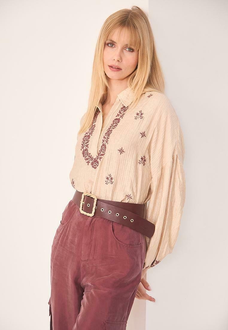 Blusa crema con bordado floral burdeos, cuello abotonado y mangas amplias, combinada con pantalones de tiro alto en un rojo profundo y un cinturón ancho.