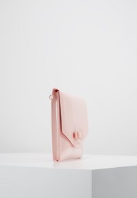 Pochette en cuir verni rose avec une fermeture à rabat et un nœud décoratif. Texture lisse et forme rectangulaire fine, posée verticalement.