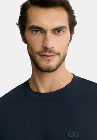 T-shirt di cotone blu navy con collo a giro, texture liscia e un piccolo logo sulla parte sinistra del petto. Inquadratura ravvicinata della parte superiore del corpo.