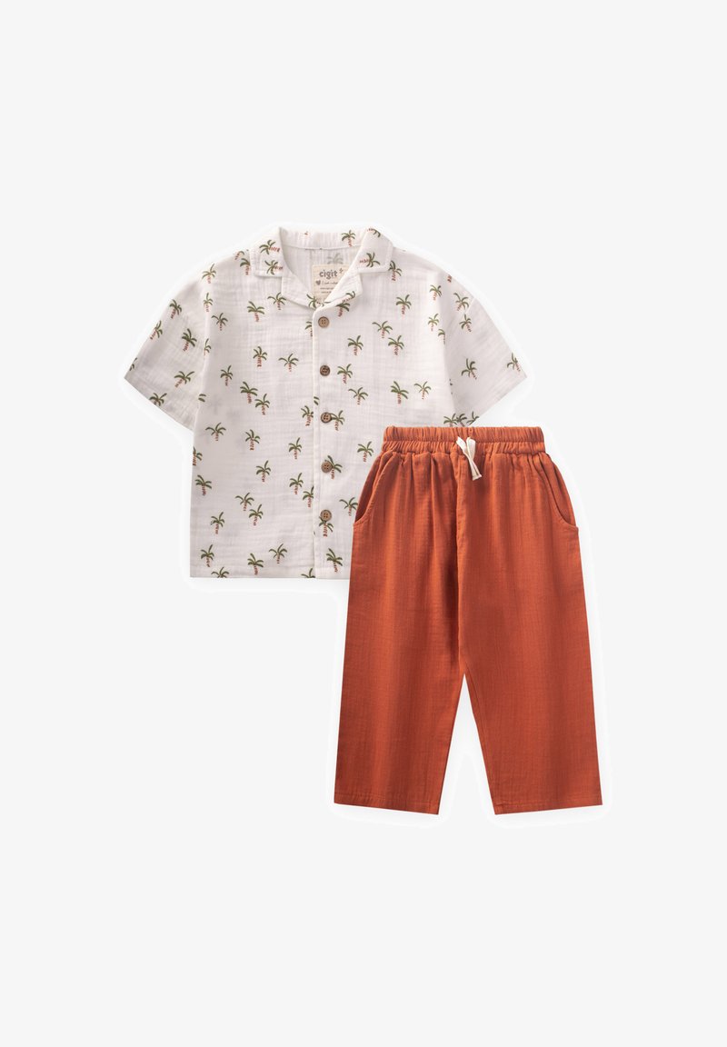 Biela košeľa s krátkymi rukávmi a zeleným potlačou palmových stromov, v kombinácii s oranžovými nohavicami na šnúrku, oboje z ľahkého materiálu.