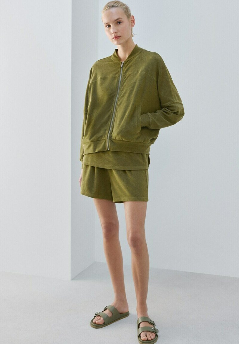 Woman wearing olive green zip-up jacket, matching shorts, and green buckle sandals, standing in a minimalist indoor space.