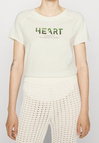 Bavlněné tričko v off-white barvě s potiskem v zelené barvě "HEART" a sloganem pod ním. Spárováno s pletenými krémovými kalhotami, které ukazují vrstvenou strukturu.