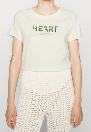 T-shirt in cotone off-white con stampa verde "HEART" e slogan sottostante. Abbinato a pantaloni a maglia aperta crema, che mostrano una texture a strati.
