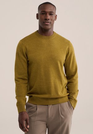 Homme portant un pull jaune moutarde à manches longues et un pantalon beige, debout avec une main dans la poche sur un fond clair uni.