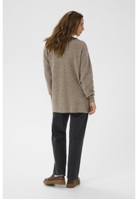 Brūns adīts džemperis ar brīvu piegriezumu, nolaižamām plecu daļām, garām piedurknēm, apvienots ar tumši pelēkiem biksēm un brūnām kurpēm.