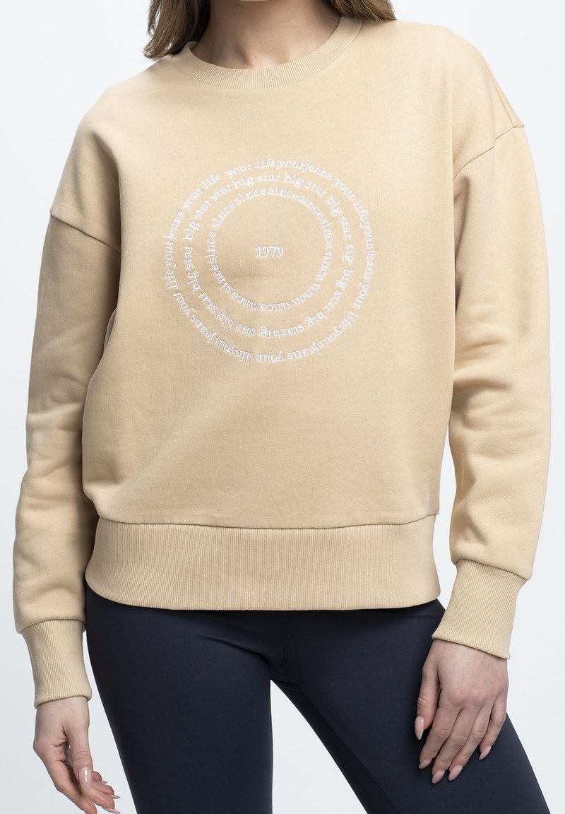 Sweat-shirt beige avec un design de texte circulaire blanc et l'année 1979 imprimée au centre, porté avec un pantalon foncé.