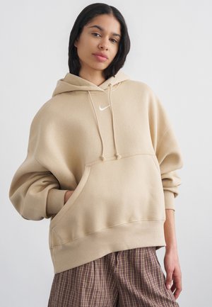 Młoda osoba z warkoczami, ubrana w beżową bluzę Nike z kapturem i kraciaste spodnie, stojąca z jedną ręką w kieszeni bluzy.