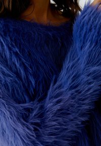 Nahaufnahme einer Person, die einen lebhaft blauen, flauschigen Pullover trägt, der die weichen, strukturierten Fasern betont, sowie einen teilweisen Blick auf den Hals und die Haare. 