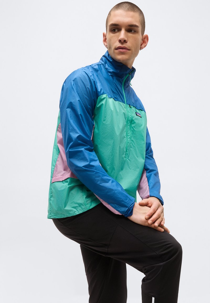 Junger Mann trägt eine blau-grün-lila Windjacke, sitzt mit den Händen über dem angehobenen Knie verschränkt und schaut zur Seite.