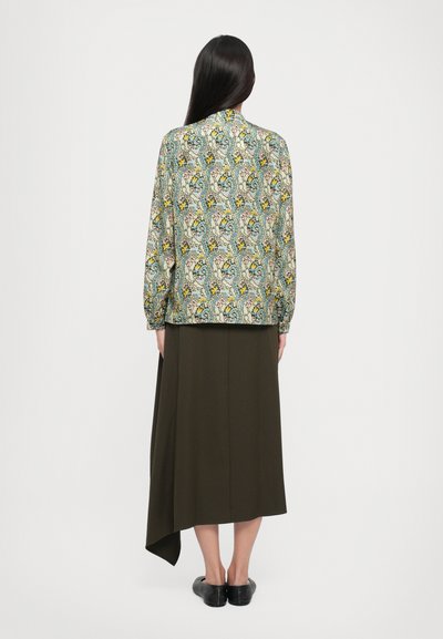 Bluză cu model floral, cu o bază de verde deschis, mâneci lungi și un croi lejer, purtată cu o fustă asimetrică de măsliniu închis.
