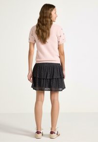 Ljusrosa kortärmad stickad tröja med broderade detaljer, kombinerad med en svart spetsfållad kjol, och beige sneakers med burgunderfärgade accenter.