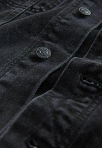 Mustast teksariidest kangas nähtava tekstuuriga; sisaldab tumedaid nööpe, õmbluste detaile ja kerget kortsus välimust. Suurendatud vaade.