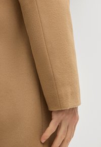 Manica di un cappotto in lana di cammello, texture liscia, vestibilità su misura, con una semplice cucitura e dettaglio a polsino arrotondato. Mano appoggiata sul tessuto.