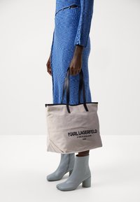 Beige canvas tote bag met zwarte leren handvatten, met een gedurfde zwarte tekstlogo. Model draagt een gestructureerde blauwe jurk en lichtblauwe enkellaarzen.