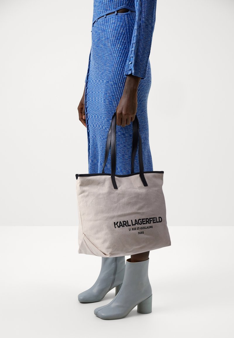 Beige canvas tote bag met zwarte leren handvatten, met een gedurfde zwarte tekstlogo. Model draagt een gestructureerde blauwe jurk en lichtblauwe enkellaarzen.