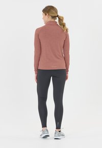 Kvinde med blond hestehale iført en rustfarvet langærmet top og sorte leggings, der står med ryggen mod kameraet på en hvid baggrund.