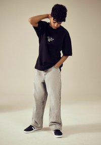 Schwarzes, übergroßes T-Shirt mit weißem "New York 92"-Logo, kombiniert mit hellgrauen, zerfetzten, weiten Jeans und schwarzen Sneakers mit weißen Akzenten.