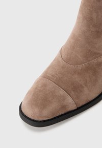 Botín de ante en marrón claro con textura suave, punta afilada y suela negra. Presenta detalles de costura en la parte superior.