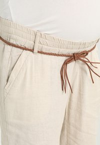 Béžové lněné kalhoty s elastickým pasem, se hnědým pleteným opaskem. Jsou viditelné boční kapsy, které zdůrazňují volnočasový design.