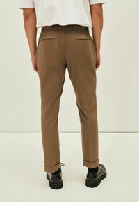 Pantaloni marroni su misura con una vestibilità slim, dotati di due tasche posteriori e orlo arrotolato. Indossati con scarpe nere.