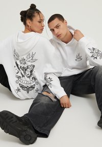 Două persoane stau pe podea, purtând hanorace albe cu modele negre în stil tatuaj de fluture, pasăre, stea și trandafir, asortate cu pantaloni negri.