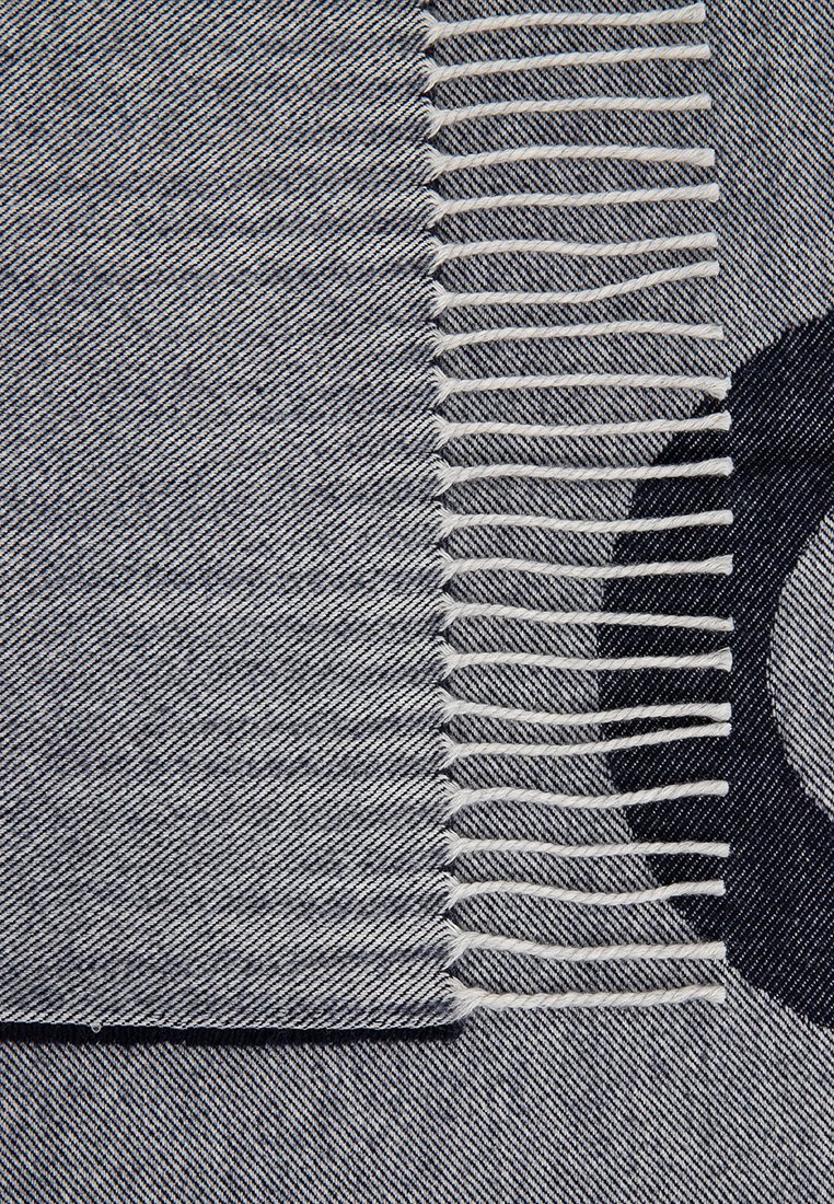 Nahaufnahme eines grauen gewebten Textils mit weißen verdrehten Fransen und einem teilweise sichtbaren schwarzen kreisförmigen Muster darunter.