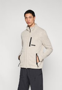 Μπεζ φλις μπουφάν με ψηλό γιακά, εμπρόσθιο φερμουάρ, μαύρες λεπτομέρειες, τσέπες με φερμουάρ και υφασμάτινη υφή. Συνδυασμένο με σκουρόχρωμο παντελόνι.