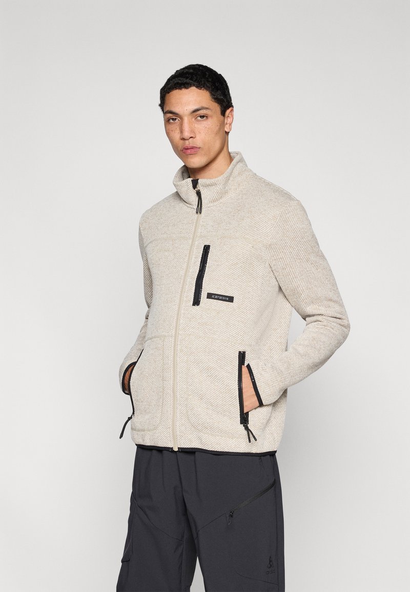 Μπεζ φλις μπουφάν με ψηλό γιακά, εμπρόσθιο φερμουάρ, μαύρες λεπτομέρειες, τσέπες με φερμουάρ και υφασμάτινη υφή. Συνδυασμένο με σκουρόχρωμο παντελόνι.