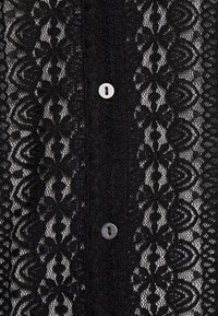 Tissu en dentelle noire présentant des motifs floraux complexes, des sections semi-transparentes et un bouton blanc au centre.