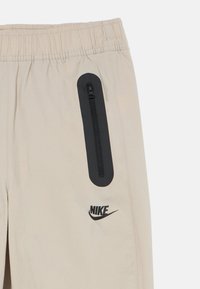 Μπεζ παντελόνια Nike με ελαστική μέση, που διαθέτουν μια μαύρη τσέπη με φερμουάρ στο πλάι και ένα μαύρο λογότυπο τυπωμένο πάνω από το στρίφωμα.