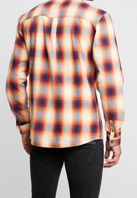 Camisa de franela a cuadros en amarillo, rojo y azul. Presenta un cuello extendido, puños con botón y un dobladillo curvado. Textura suave, mangas largas.