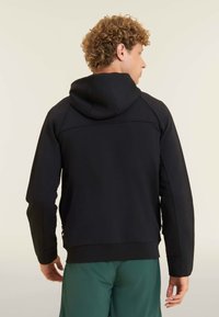 Sudadera con capucha negra de textura suave, con mangas raglán y un bolsillo con cremallera, llevada sobre pantalones cortos verdes.
