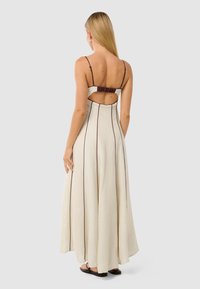 Rochie maxi crem cu dungi verticale subțiri de culoare maro, spate deschis cu detalii plisate și bretele reglabile. Material fin și siluetă fluidă.