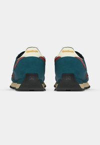 Ténis de camurça verde-água e vermelha com uma sola bege, apresentando um calcanhar em borracha preta texturizada e uma marcação amarela na parte de trás.