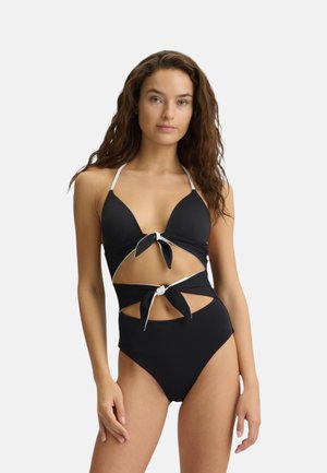 Donna con capelli lunghi e castani che indossa un costume da bagno nero con ritagli bianchi e due dettagli con fiocchi frontali, in piedi davanti a uno sfondo chiaro e uniforme.