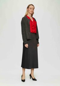Grauer Strickcardigan über einer roten Rüschbluse getragen, kombiniert mit einem schwarzen knielangen Rock und schwarzen High Heels.